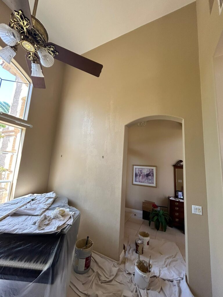 Room with tarps and paint supplies on the floor, wall partially painted beige, ceiling fan above.